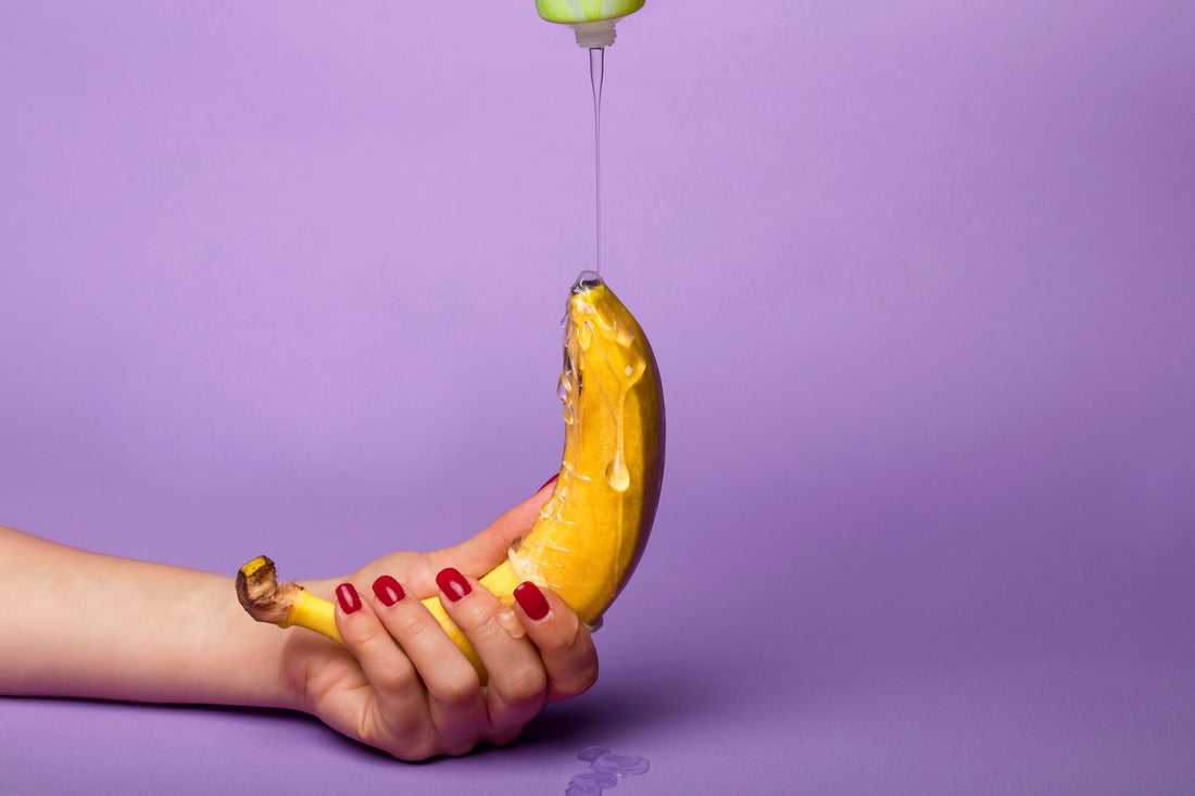 A hand holding a banana beneath a stream of clear lubricant against a purple background